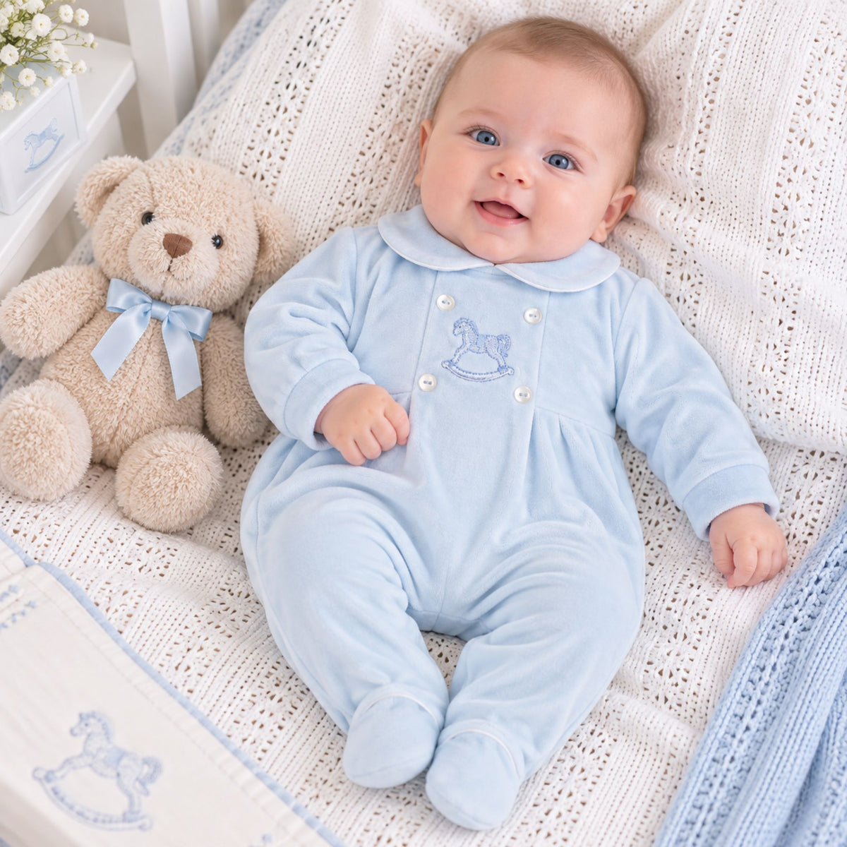 A baby wearing a light blue footed romper with a Peter Pan collar and embroidered rocking horse detail, lying on a white knit blanket next to a teddy bear.
