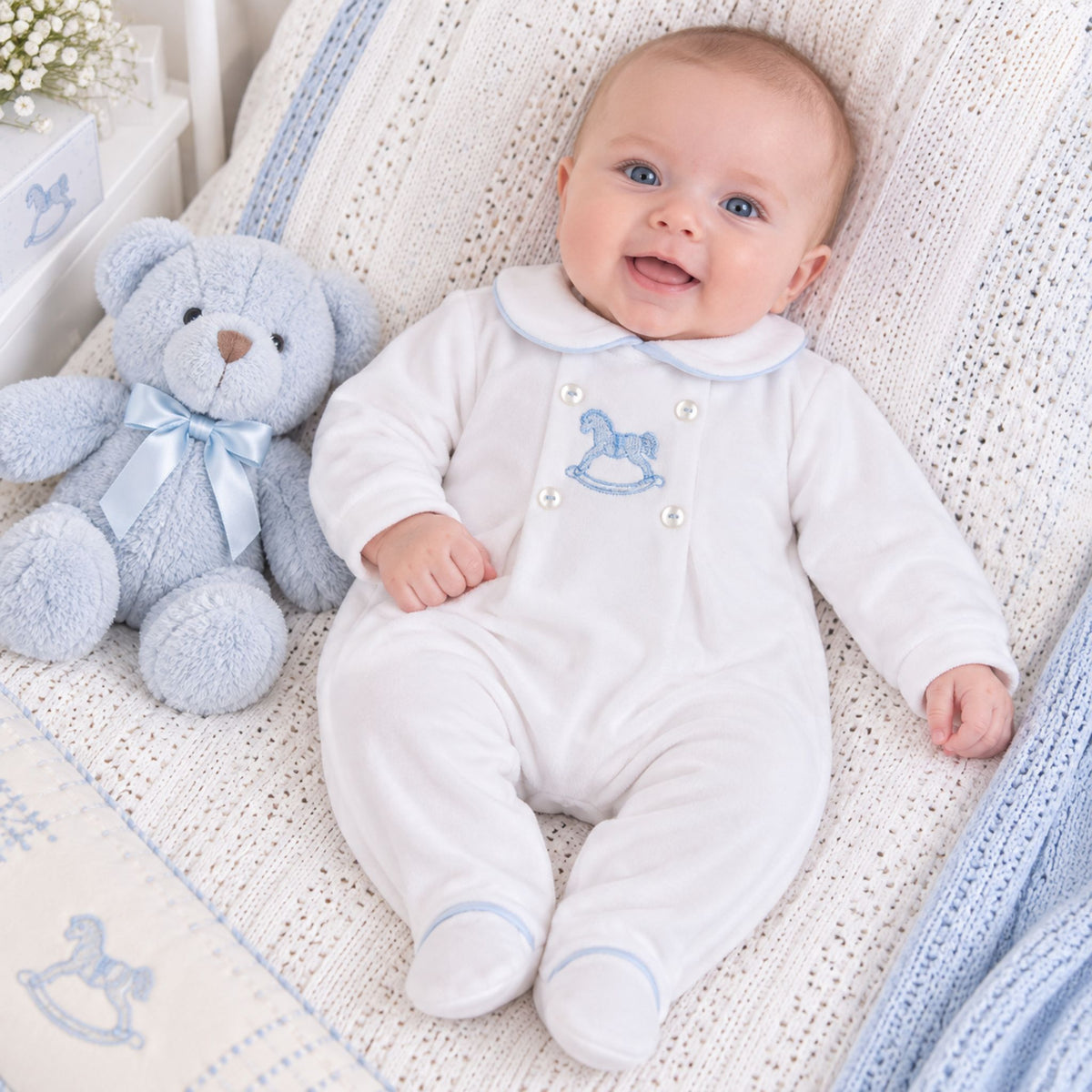 A baby wearing a white footed onesie with a blue rocking horse embroidery and pearl buttons, lying on a white knit blanket next to a light blue teddy bear.