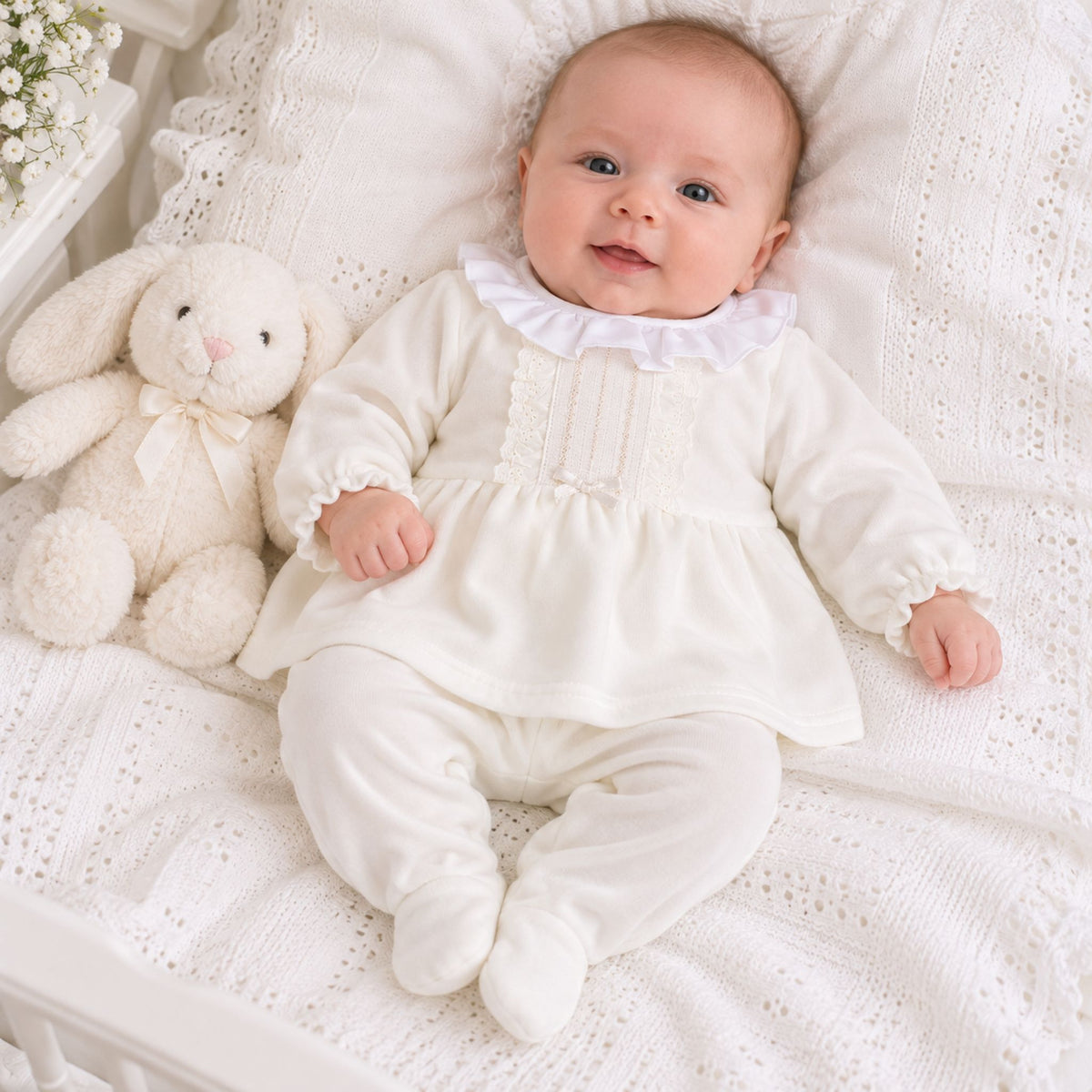 A baby wearing a cream-colored long-sleeve footed romper with a ruffled collar, lying on a white textured blanket next to a plush white bunny toy.