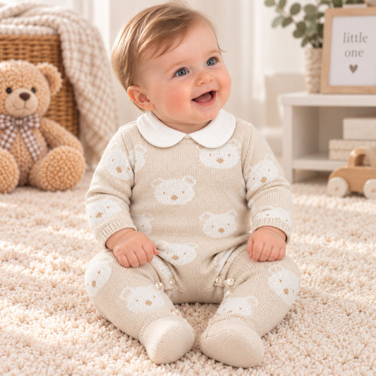 A baby sitting on a cream carpet wearing a beige knit footed romper with a white Peter Pan collar and white teddy bear prints. The background shows a cozy nursery setting with a teddy bear, a basket, and a small wooden toy cart.
