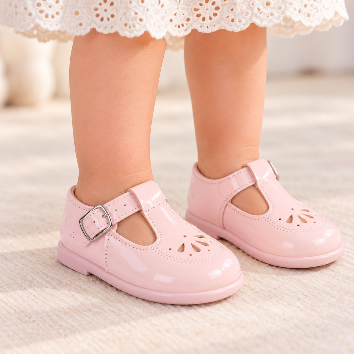 A close-up of a child's feet wearing shiny light pink Mary Jane-style shoes with cutout details and silver buckle straps, standing on a light cream textured floor.