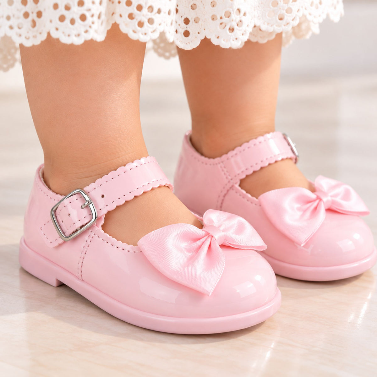 A close-up of a child's feet wearing light pink Mary Jane style shoes with satin bows and scalloped straps, standing on a light marble floor. The child is wearing a white lace skirt.