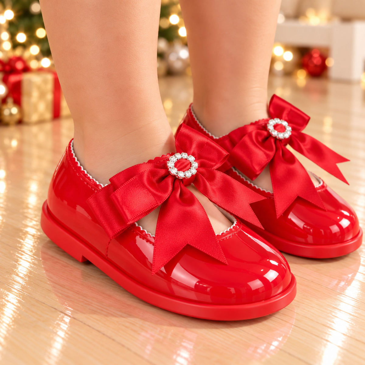 A pair of glossy red children's Mary Jane shoes with large satin bows and jeweled bow centers, worn on a child's feet in a festive indoor setting with blurred Christmas lights in the background.