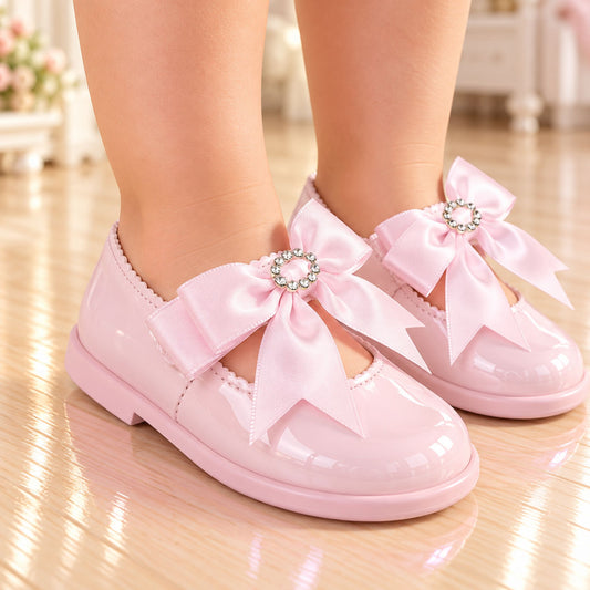 A pair of children's glossy pink patent Mary Jane shoes with large satin bows and jeweled center accents, shown being worn on a child's feet in a softly lit indoor setting.