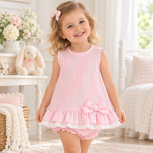A young girl with blonde hair and a pink bow smiles while wearing a pink and white vertically striped sleeveless dress with a ruffled hem and matching bloomers in a bright, decorated nursery.