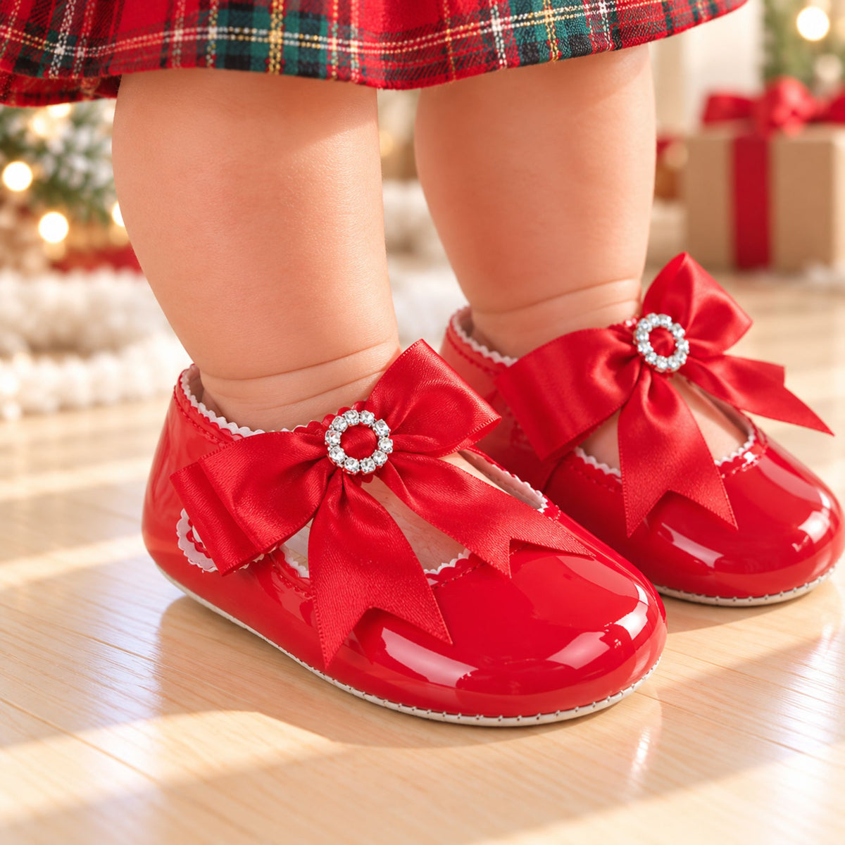 A close-up of a baby's legs wearing shiny red patent Mary Jane shoes with large red bows and jeweled centers, standing on a light wood floor in a softly blurred holiday setting.
