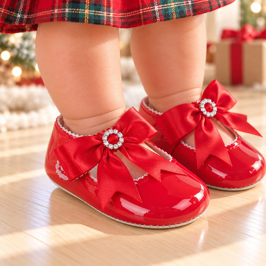 A close-up of a baby's legs wearing shiny red patent Mary Jane shoes with large red bows and jeweled centers, standing on a light wood floor in a softly blurred holiday setting.