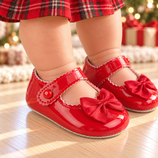 A close-up of a baby's feet wearing shiny red Mary Jane baby shoes with large bows and scalloped trim, standing on a light wood floor with a blurred Christmas background.