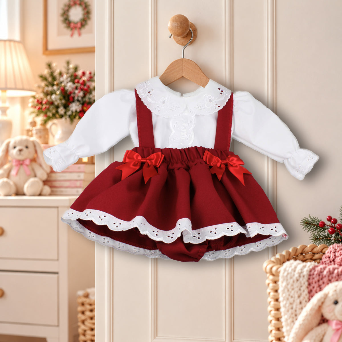 A toddler girl's holiday dress hanging on a wooden hanger against a beige paneled door. The dress features a white long-sleeve blouse with a lace collar, a deep red pinafore-style skirt with red bows, and white eyelet lace trim.