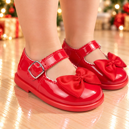 A pair of bright red patent leather Mary Jane shoes with satin bows and silver buckle straps, shown on a child's feet on a wooden floor with a blurred Christmas background.