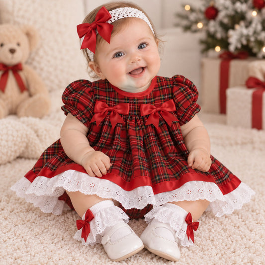 A baby girl sitting on a cream carpet wearing a red and green plaid holiday dress with white lace trim, a red bow headband, and white shoes with red bows. A teddy bear and a decorated Christmas tree are in the background.