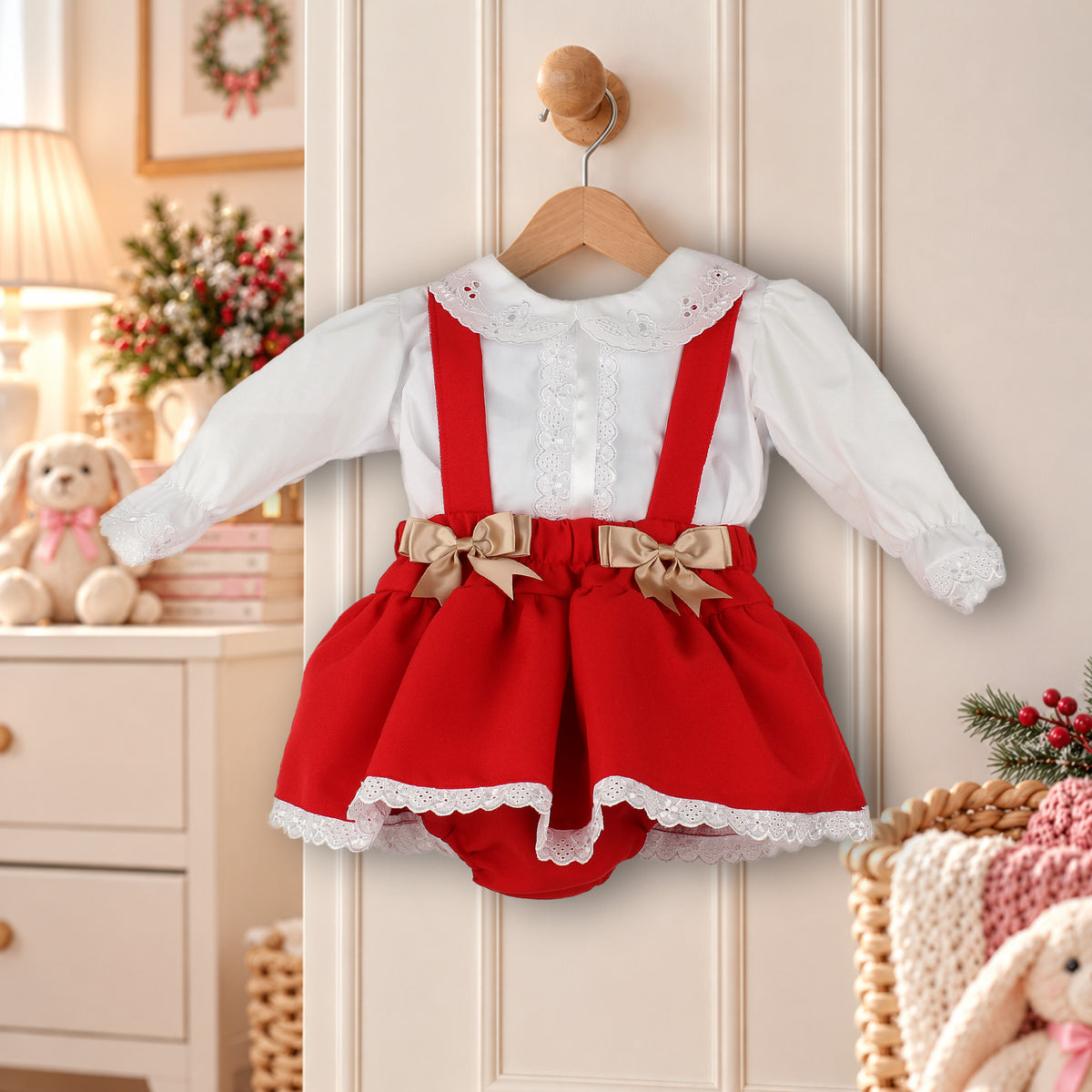 A baby girl's holiday-themed two-piece outfit consisting of a white long-sleeve blouse with lace trim and red pinafore-style overalls with beige bows and white lace hem, hanging on a wooden hanger against a cream paneled door.
