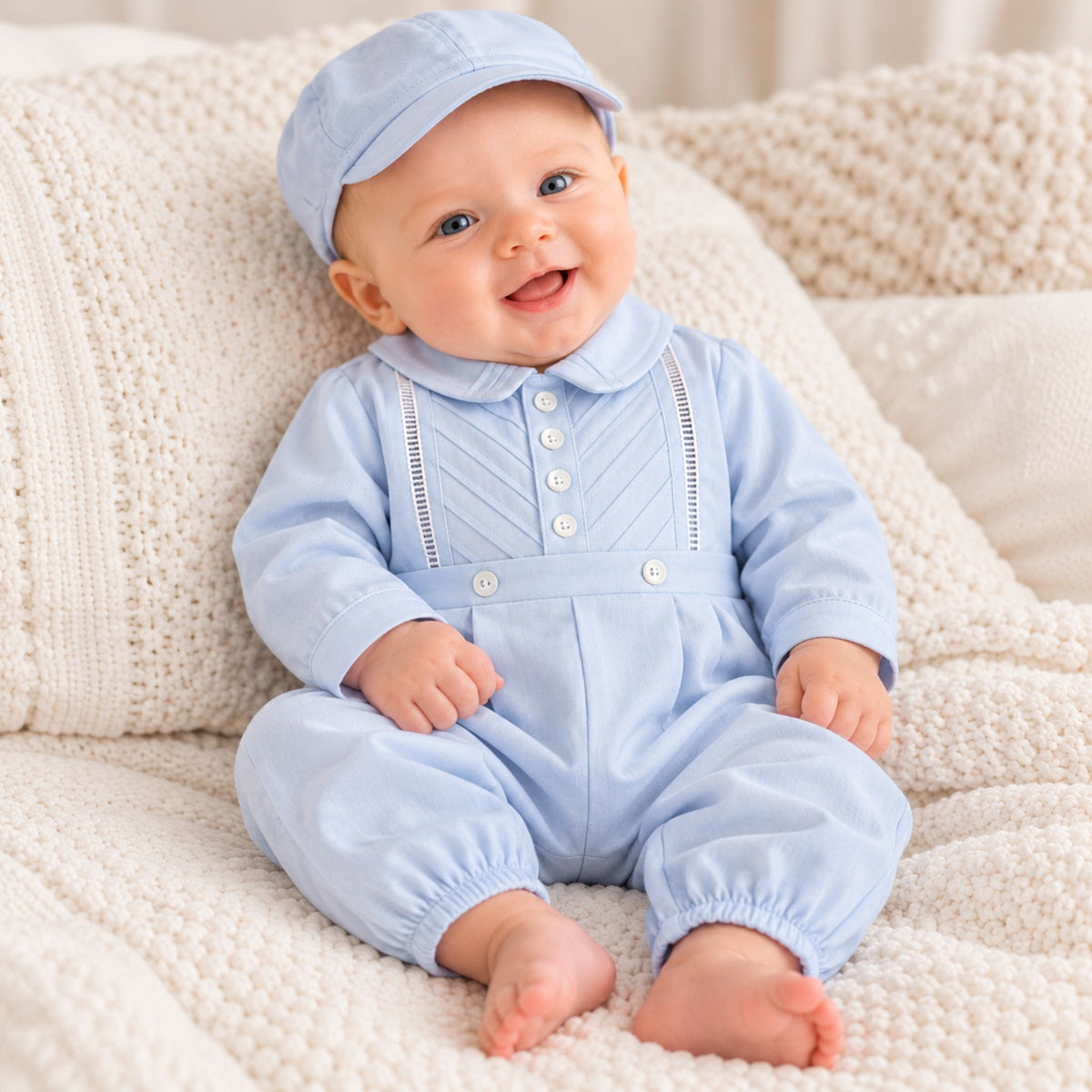 A smiling baby wearing a light blue long-sleeve romper and matching cap, sitting on a cream knit blanket.