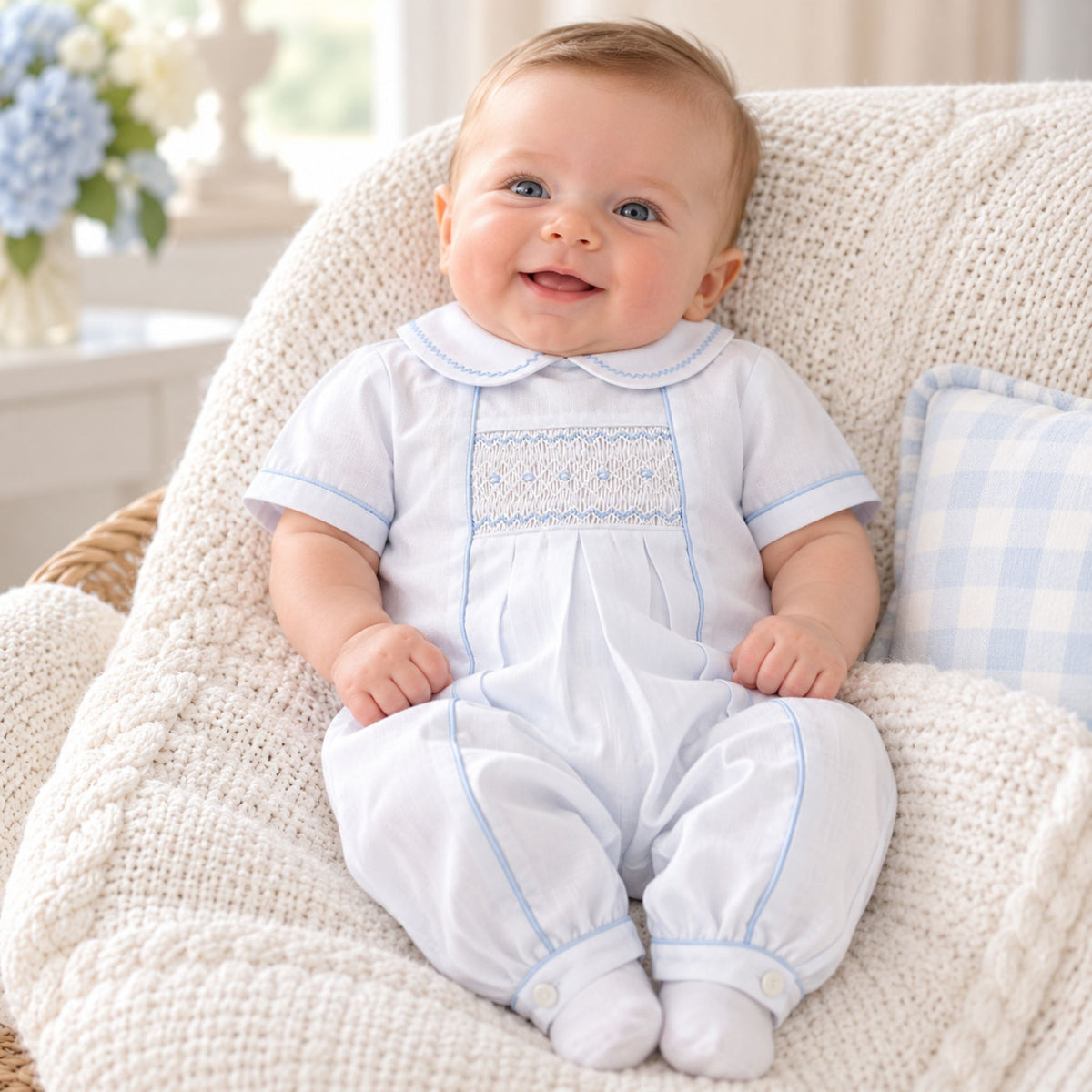 A baby sitting in a cream-colored knit armchair, wearing a white short-sleeve baby romper with light blue trim and a smocked chest panel.