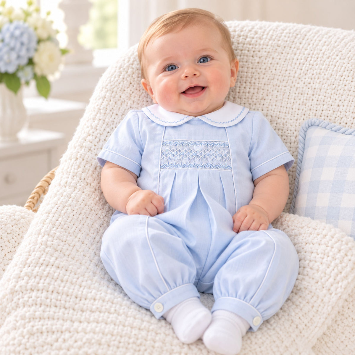 A baby sitting in a cream-colored woven chair, wearing a light blue short-sleeve baby jumpsuit with white trim and a smocked chest panel, along with white socks.