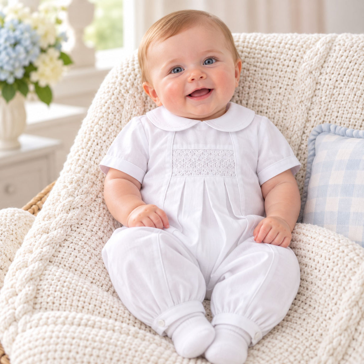 A baby wearing a white short-sleeve smocked romper with a Peter Pan collar, sitting on a cream-colored knit chair.