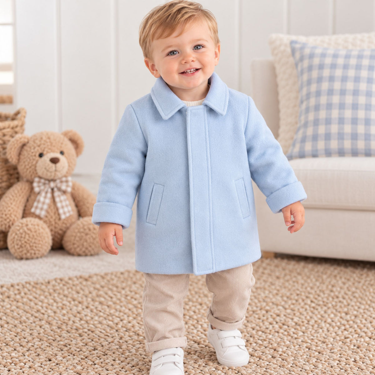 A toddler wearing a light blue coat, beige pants, and white shoes stands on a textured rug in a bright room with a teddy bear and a sofa in the background.