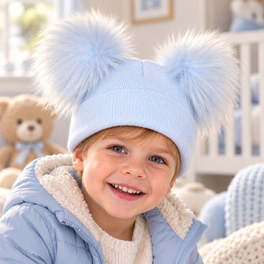 A smiling young child wearing a light blue knit beanie with two large fluffy pom-poms, photographed indoors in a bright nursery-like setting.