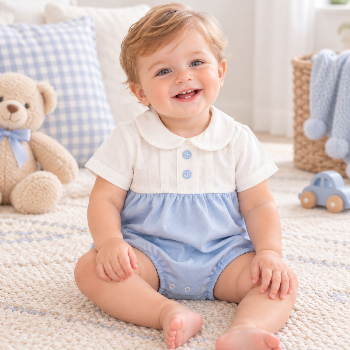 A smiling baby boy sitting on a textured cream rug, wearing a short-sleeve baby romper with a white top and light blue bottom. The background shows a cozy nursery setting with a blue and white gingham pillow, a beige teddy bear, and a small toy car.