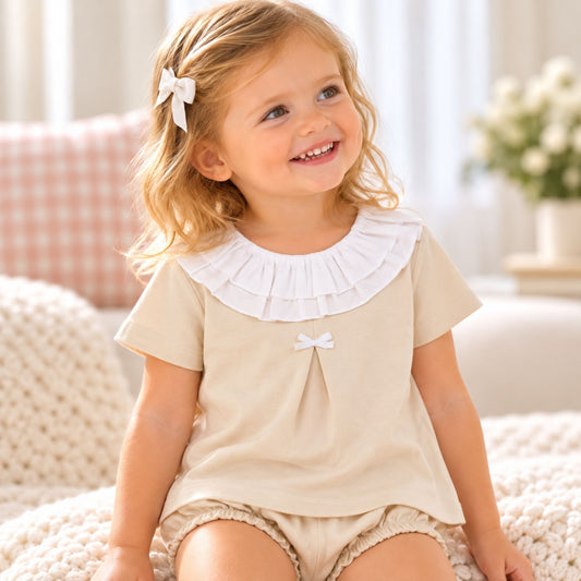 A young girl with blonde hair wearing a beige short-sleeve dress with a white ruffled collar and matching beige shorts, sitting on a cozy white knit blanket in a bright room.