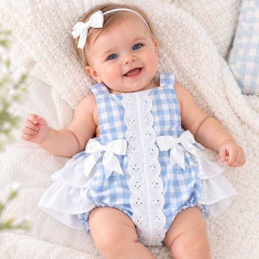 A baby wearing a light blue and white gingham romper with a white eyelet center panel, white bow details, and a white ruffled hem, lying on a soft cream blanket.