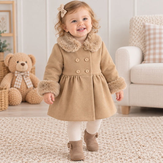 A toddler girl stands in a cozy room wearing a beige wool-like coat with a faux-fur collar and cuffs, white tights, and brown ankle boots. A teddy bear sits on the left, and a light-colored armchair is on the right.