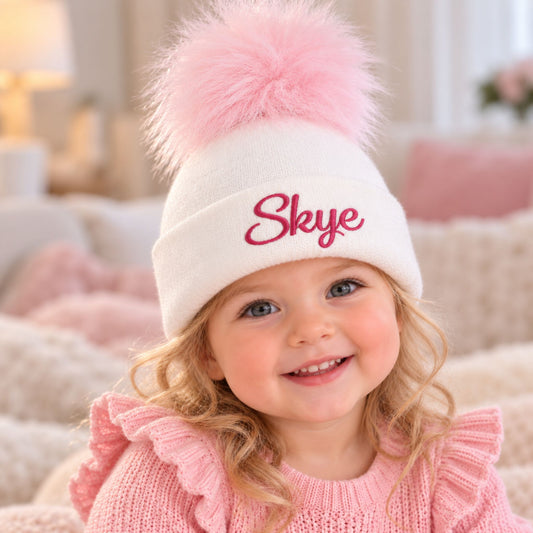 A smiling young girl wearing a white knit pom-pom beanie with the name 'Skye' embroidered in pink on the front, paired with a pink knit sweater in a cozy, softly lit room.