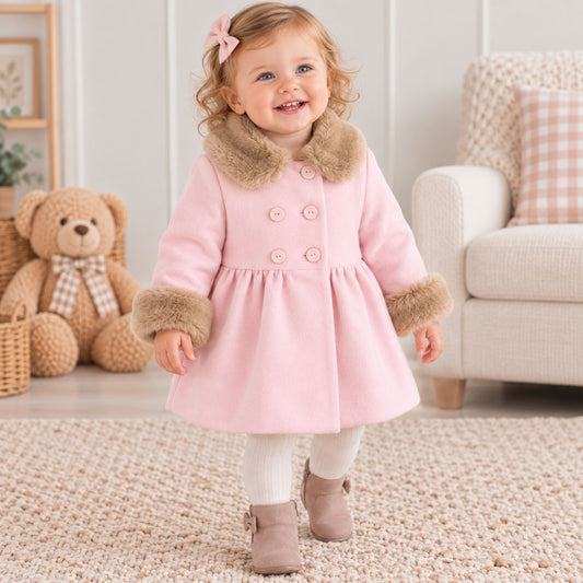 A toddler girl wearing a light pink winter coat with brown faux-fur trim, white leggings, and beige boots, standing on a textured rug in a cozy room with a teddy bear and sofa in the background.