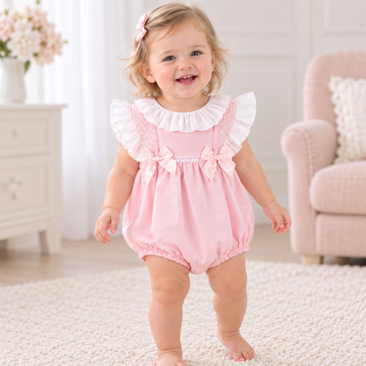 A toddler girl wearing a pink romper with white ruffle trim and bow details, standing on a white rug in a bright, neutral-toned room.