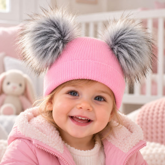 A smiling toddler wearing a pink knit beanie with two large gray faux-fur pom-poms, paired with a pink jacket in a soft pink nursery setting.