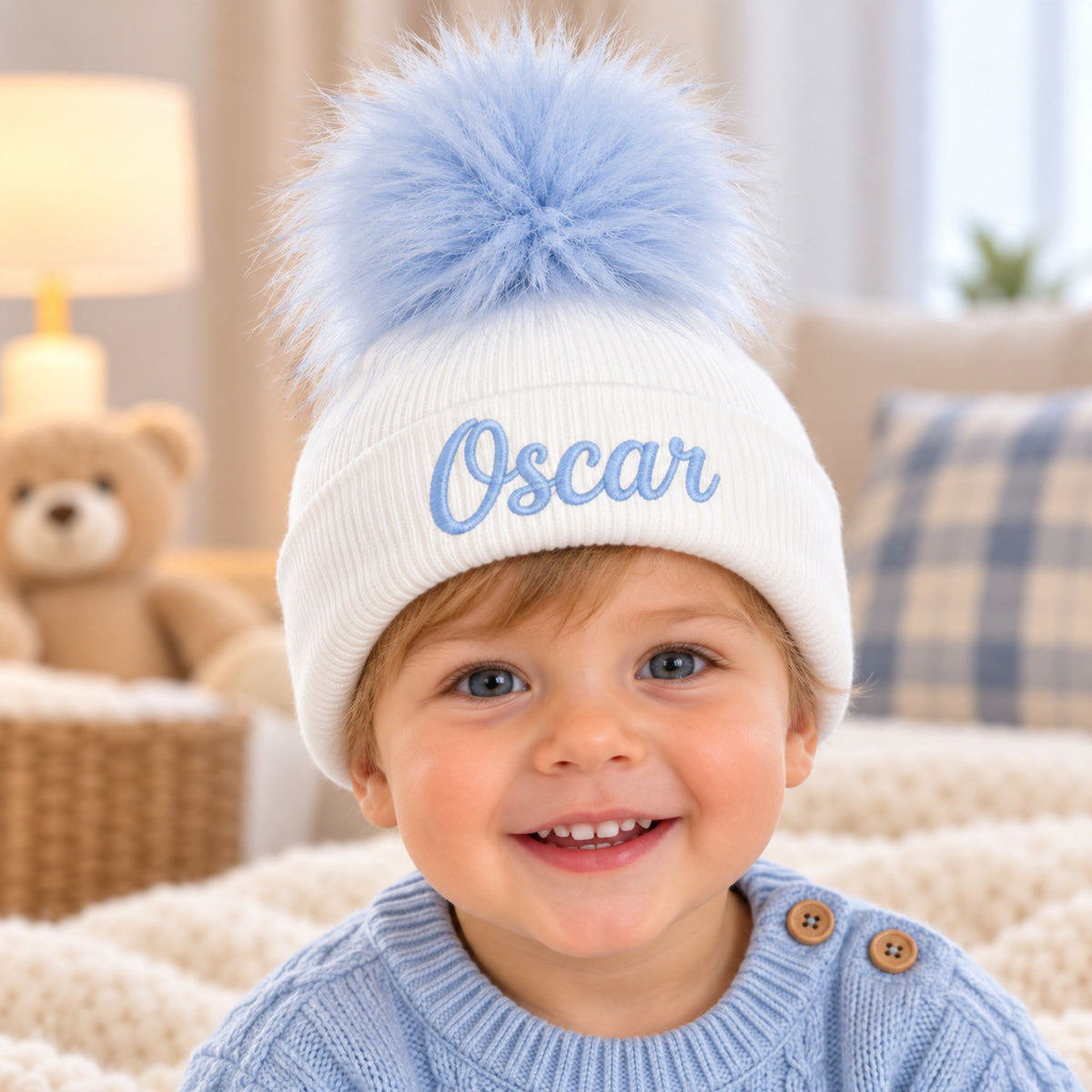 A smiling toddler wearing a white knit pom-pom beanie with the name 'Oscar' embroidered in light blue on the front, posed in a cozy indoor setting.
