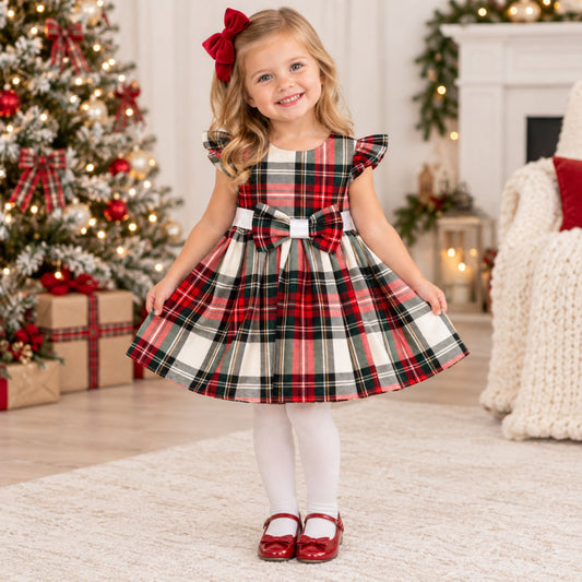 A young girl stands in a decorated holiday living room wearing a red, green, and white plaid dress with ruffled sleeves, white tights, red shoes, and a red bow in her hair.