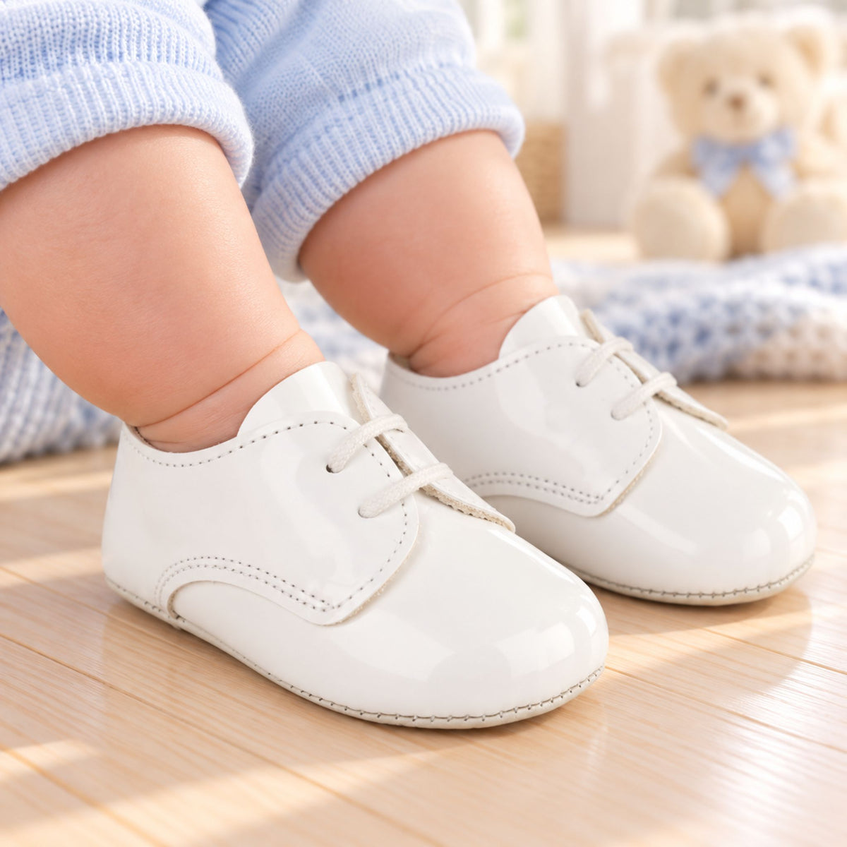 A baby wearing shiny white lace-up baby shoes and light blue knit pants, standing on a light wooden floor with a soft-focus teddy bear in the background.
