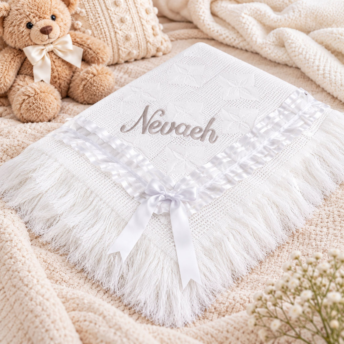 A white personalized baby shawl with fringe trim, a satin ribbon bow, and the name 'Nevaeh' embroidered on the front, styled on a beige knit blanket with a teddy bear and small flowers.