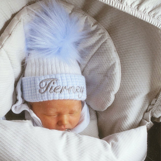A newborn baby wearing a white and blue checkered bobble hat with a blue pom pom and personalized name 'Dylan' in silver.
